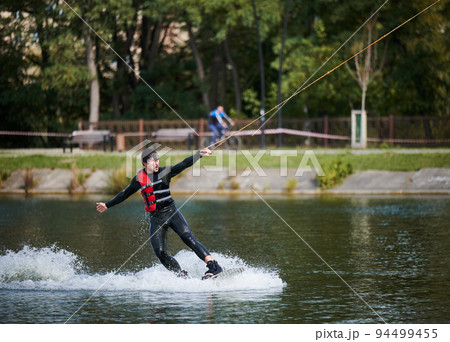 Wakeboarder surfing on lake. Young man surfer having fun wakeboarding in the cable park. Water sport, outdoor activity concept. 94499455