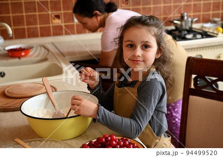 Beautiful Caucasian child, adorable little little girl helps her mother in kitchen, learns culinary, kneads dough, prepares cherry pie, smiles at camera 94499520