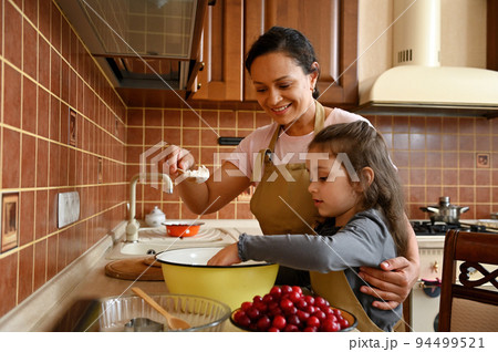 Beautiful African American young woman in chef apron, loving mom smiling and hugging her daughter, adorable multi-ethnic little child girl, while teaching her cooking homemade pie in the home kitchen Beautiful African American young woman in chef apron, loving mom smiling and hugging her daughter, adorable multi-ethnic little child girl, while teaching her cooking homemade pie in the home kitchen 94499521