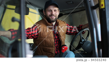Portrait of caucasian male farmer in cap sitting in tractor with open door and smiling to camera. Agriculture farming. 94503240