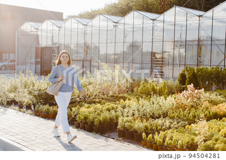 A young pretty woman walks down the street near the large greenhouses. A woman goes to buy green plants for decoration. Landscaping concept 94504281