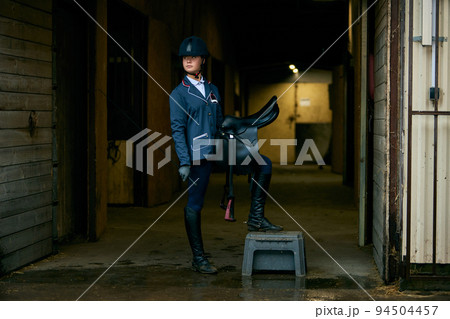 Portrait of young girl, jockey and horsewoman in competition uniform and helmet standing stands in stable before the start of horse race Portrait of young girl, jockey and horsewoman in competition uniform and helmet standing stands in stable before the start of horse race 94504457