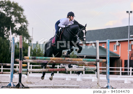 Jump over barrier. Young sportive girl, teen training at riding arena in summer day, outdoors. Dressage of horses. Horseback riding. Model in sports uniform 94504470