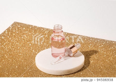Pink glass bottle of cosmetic liquid transparent gel on white background with golden sheet. Dropper bottle, hyaluronic acid, oil, serum, skin care product. Copy space. 94506938