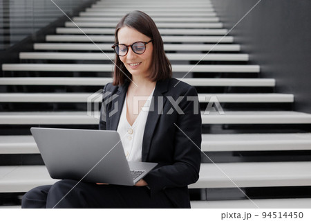 business woman in a formal suit with a laptop in an office or business center sits on the steps in the lobby and works online business woman in a formal suit with a laptop in an office or business center sits on the steps in the lobby and works online 94514450
