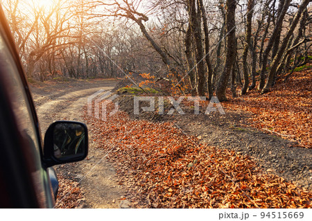Side rear mirror view of SUV car drive on beautiful dirt gravel forest unpaved road in autumn foliage and bright warm sunlight through woods background. Off road car adventure nature fall journey Side rear mirror view of SUV car drive on beautiful dirt gravel forest unpaved road in autumn foliage and bright warm sunlight through woods background. Off road car adventure nature fall journey 94515669