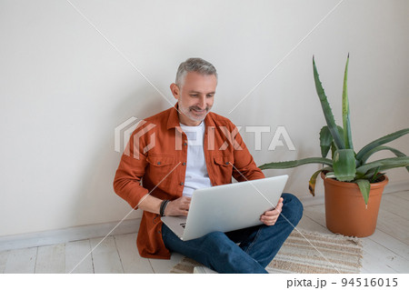 Man in terracotta shirt sitting on the floor with a laptop in hands Man in terracotta shirt sitting on the floor with a laptop in hands 94516015