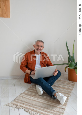 Man in terracotta shirt sitting on the floor with a laptop in hands 94516016