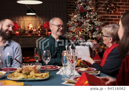 Family celebrating christmas, exchanging gifts at festive served table, eating traditional dishes at home feast. Xmas celebration with parents, people giving present boxes 94516062