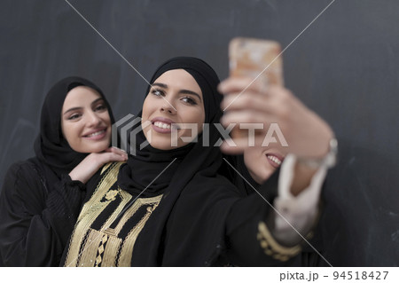 Group of young beautiful muslim women in fashionable dress with hijab using smartphone while taking selfie picture in front of black background 94518427