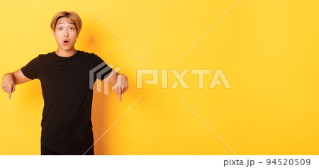 Amused happy asian guy, pointing fingers down and gasping impressed, showing awesome advertisement, yellow background 94520509