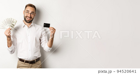Image of young businessman holding credit card and money, looking at upper left corner and thinking about shopping, standing over white background Image of young businessman holding credit card and money, looking at upper left corner and thinking about shopping, standing over white background 94520641