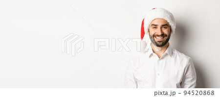 Close-up of happy bearded man celebrating christmas, wearing santa party hat and smiling, standing over white background 94520868