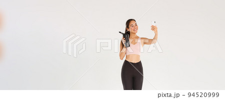 Full length of smiling slim and healthy asian girl taking selfie fitness training session, female blogger and athelte showing water bottle on camera of mobile phone, standing over white background Full length of smiling slim and healthy asian girl taking selfie fitness training session, female blogger and athelte showing water bottle on camera of mobile phone, standing over white background 94520999