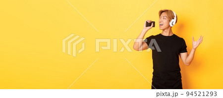 Joyful attractive korean guy in headphones, playing karaoke app, singing into mobile phone microphone, standing yellow background Joyful attractive korean guy in headphones, playing karaoke app, singing into mobile phone microphone, standing yellow background 94521293