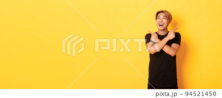 Portrait of happy smiling asian man with blond hair, looking amused left, pointing sideways at two variants, yellow background Portrait of happy smiling asian man with blond hair, looking amused left, pointing sideways at two variants, yellow background 94521450