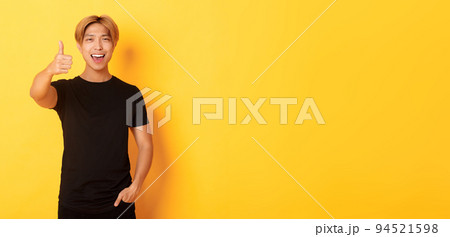Portrait of pleased handsome asian guy, showing thumbs-up in approval, standing over yellow background 94521598