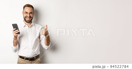 Satisfied business man showing thumbs up after using mobile phone, standing pleased over white background 94522784