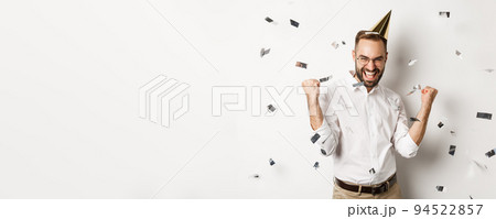 Celebration and holidays. Happy man dancing on birthday party with confetti, wearing b-day hat and rejoicing, standing over white background Celebration and holidays. Happy man dancing on birthday party with confetti, wearing b-day hat and rejoicing, standing over white background 94522857