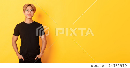 Handsome asian blond guy in glasses standing in black clothes and smiling pleased, yellow background 94522959