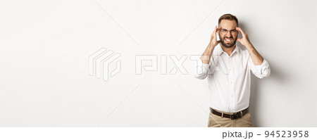 Businessman having headache, grimacing and holding hands on head, standing over white background Businessman having headache, grimacing and holding hands on head, standing over white background 94523958