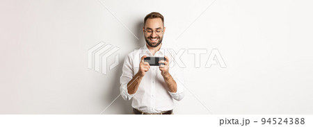 Happy businessman watching video on mobile phone, standing over white background Happy businessman watching video on mobile phone, standing over white background 94524388