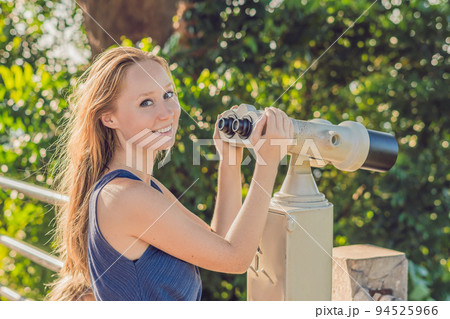 young beautiful blond woman enjoy the view with an coin operated binoculars. The water and the sky is blue. she wears a white dress and sunglasses. she feels good, is smiling and look to the horizon 94525966