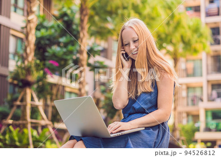 Young female freelancer sitting near the pool with her laptop in the hotel browsing in her smartphone. Busy at holidays. Distant work concept. Copy space for your text 94526812