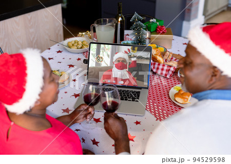 African american senior couple with vine on christmas video call with santa claus in face maks 94529598