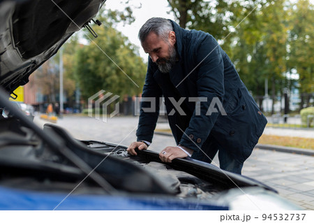 mature man trying to figure out a car breakdown leaning over the engine in the city 94532737