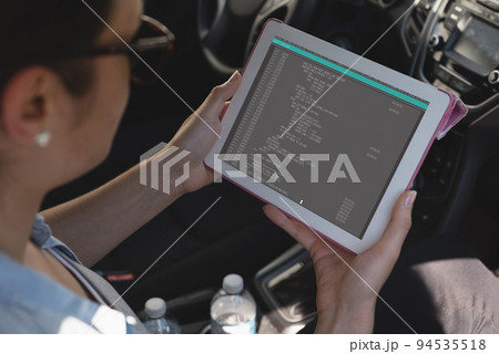 Biracial female programmer sitting in car, using tablet with coding on screen 94535518