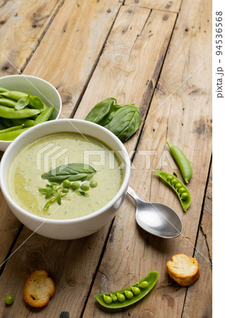 Vertical image of bowl of pea soup, with peas and basil leaves on wood, with copy space 94536568