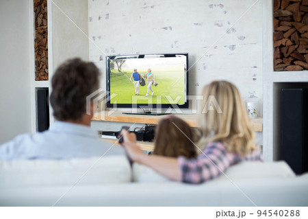 Rear view of family sitting at home together watching golf event on tv Rear view of family sitting at home together watching golf event on tv 94540288