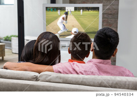 Rear view of family sitting at home together watching cricket match on tv Rear view of family sitting at home together watching cricket match on tv 94540334