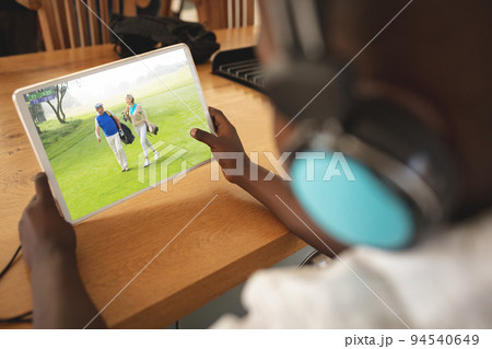 African american boy wearing headphones at home watching senior couple playing golf on tablet African american boy wearing headphones at home watching senior couple playing golf on tablet 94540649