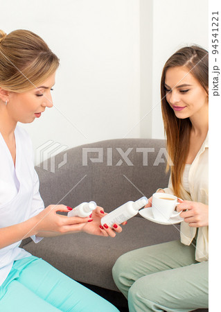 Beautician offering product for the young woman holding white plastic bottles with a cream sitting on the sofa Beautician offering product for the young woman holding white plastic bottles with a cream sitting on the sofa 94541221