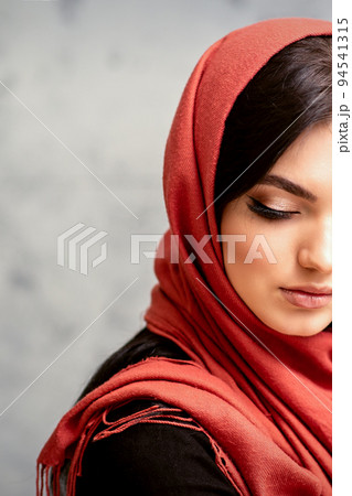 The fashionable young woman. Portrait of the beautiful female model with long hair and makeup with eyelash extensions in a red scarf. Beauty young caucasian woman on the background of a gray wall The fashionable young woman. Portrait of the beautiful female model with long hair and makeup with eyelash extensions in a red scarf. Beauty young caucasian woman on the background of a gray wall 94541315