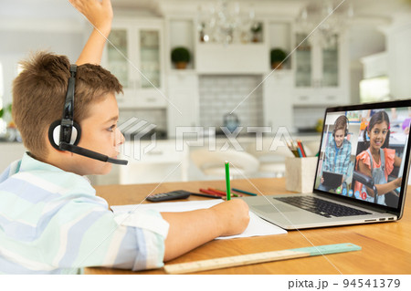 Caucasian boy raising hand for video call, with smiling diverse elementary school pupils on screen 94541379