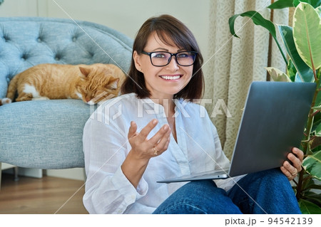 Middle aged woman using laptop, with sleeping cat lying on armchair 94542139
