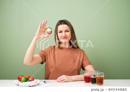 A young beautiful woman in a brown t-shirt paints Easter eggs in different colors. Preparing Easter Eve at home. Green wall. 94544880