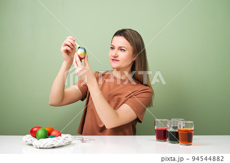 A young beautiful woman in a brown t-shirt paints Easter eggs in different colors. Preparing Easter Eve at home. Green wall. A young beautiful woman in a brown t-shirt paints Easter eggs in different colors. Preparing Easter Eve at home. Green wall. 94544882