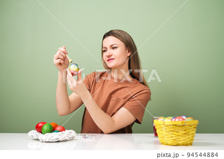 A young beautiful woman in a brown t-shirt paints Easter eggs in different colors. Preparing Easter Eve at home. Green wall. A young beautiful woman in a brown t-shirt paints Easter eggs in different colors. Preparing Easter Eve at home. Green wall. 94544884