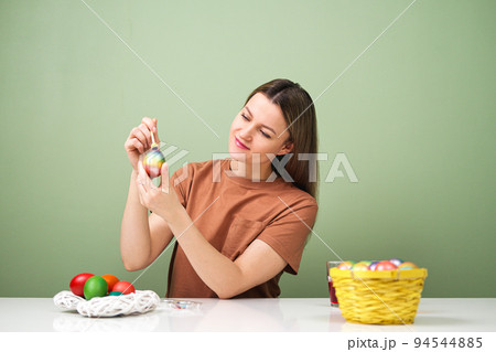 A young beautiful woman in a brown t-shirt paints Easter eggs in different colors. Preparing Easter Eve at home. Green wall. A young beautiful woman in a brown t-shirt paints Easter eggs in different colors. Preparing Easter Eve at home. Green wall. 94544885