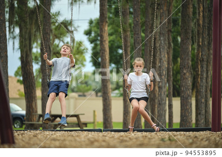 Two young teenage children, girl and boy playing on swings together outdoors on bright sunny vacations day 94553895