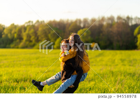 Playful mother giving daughter piggy back ride at green field. 94558978