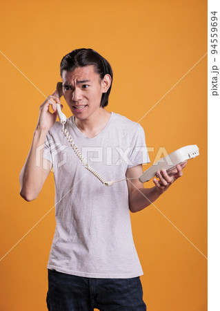 Young angry man talking on landline phone. Irritated asian teenager shouting, holding retro telephone, person with annoyed facial expression having conversation, answering call Young angry man talking on landline phone. Irritated asian teenager shouting, holding retro telephone, person with annoyed facial expression having conversation, answering call 94559694