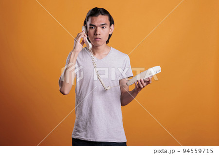 Young scared man talking on landline phone. Confused asian teen chatting, holding retro telephone, frightened person with terrified facial expression answering call on orange background 94559715