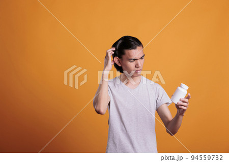 Asian young man confused about what pills to take, looking at tablets bottle. Disease treatment, illness remedy, upset puzzled patient holding medicaments, reading instruction 94559732