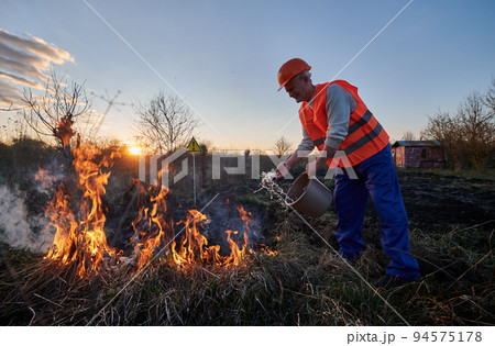Fireman ecologist extinguishing fire in field with warning sign with exclamation mark end evening sky on background. Male environmentalist holding bucket and pouring water on burning dry grass. Fireman ecologist extinguishing fire in field with warning sign with exclamation mark end evening sky on background. Male environmentalist holding bucket and pouring water on burning dry grass. 94575178
