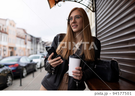 young woman with a cup of coffee by a mobile phone with a smile looks into the distance against the young woman with a cup of coffee by a mobile phone with a smile looks into the distance against the 94577501
